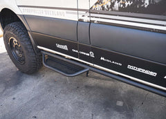 Driver side view looking from the rear toward the front, showcasing the full ROAMBUILT Mercedes Sprinter Side Step mounted under the van with visible perforated aluminum decking 