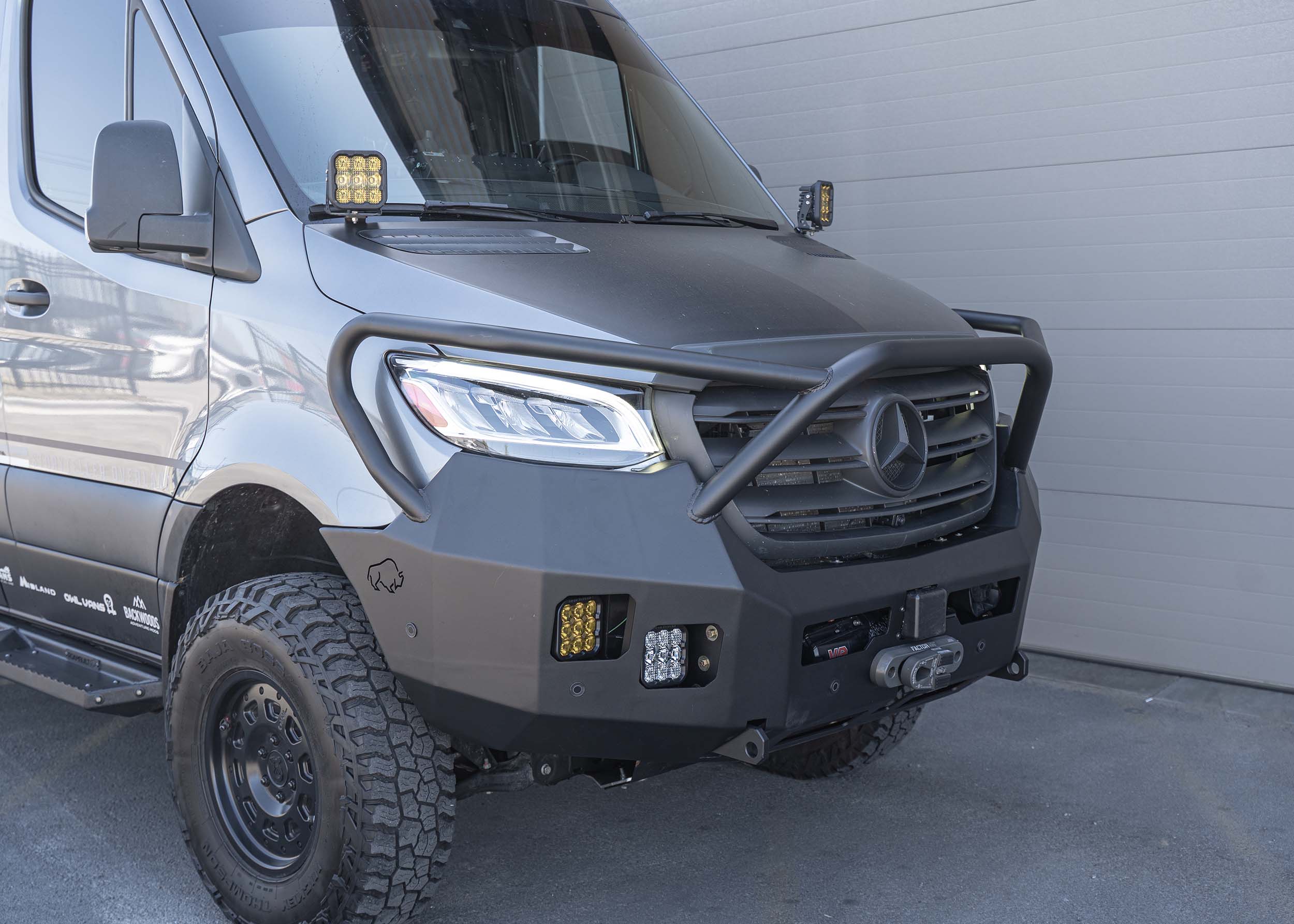 Passenger-side view of a Mercedes Sprinter van featuring the Full Guard Front Bumper with a hybrid steel and aluminum construction, showcasing its full front-end protection, integrated bars, front-mounted winch cradle, and all designed for lightweight durability and rugged off-road performance.