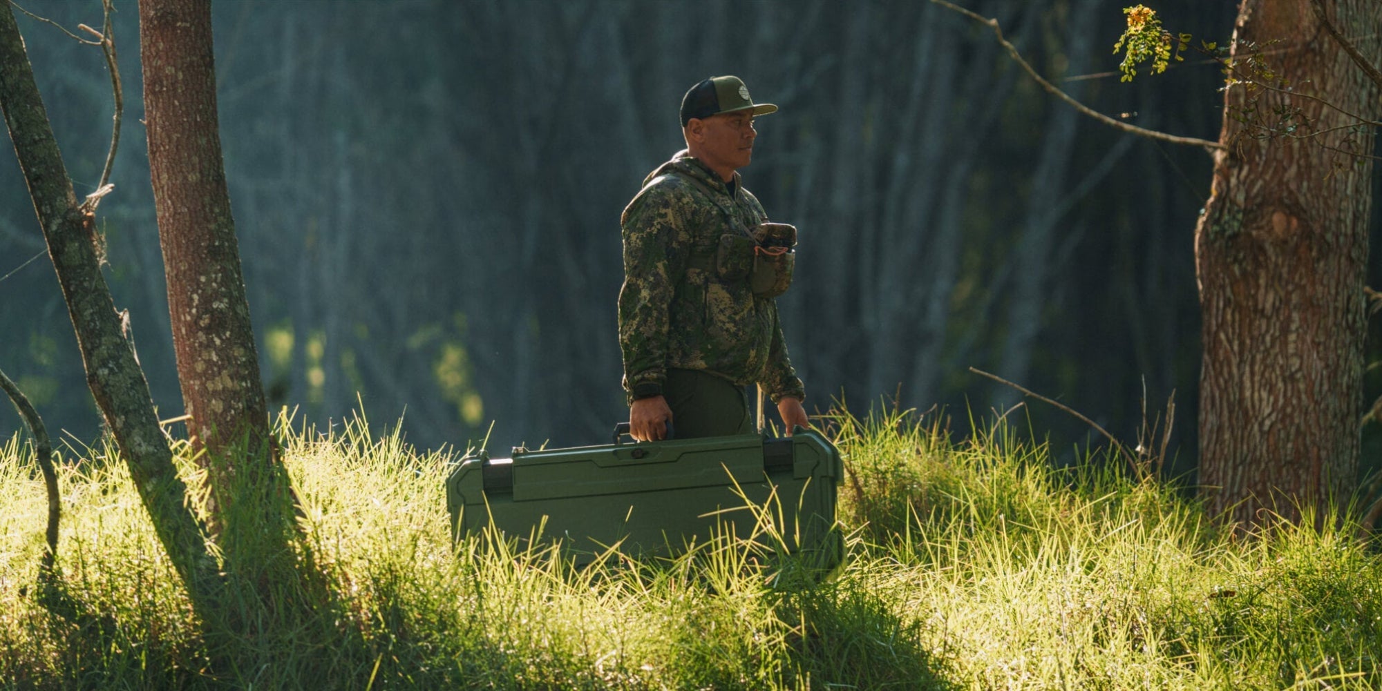 [A0031-MANF-GRN] @expanded A man in camo walking through the woods, carrying his Ranger Green Honcho 80 D-co case that he uses for his bow.